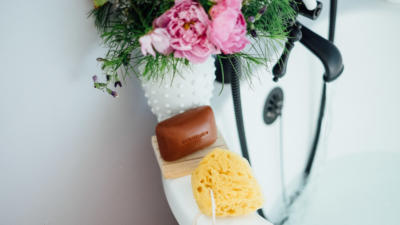 Schmidt's Rose+Vanilla bar soap on the side of a white tub with a loofah and pink floral arrangement as the tub fills up with water